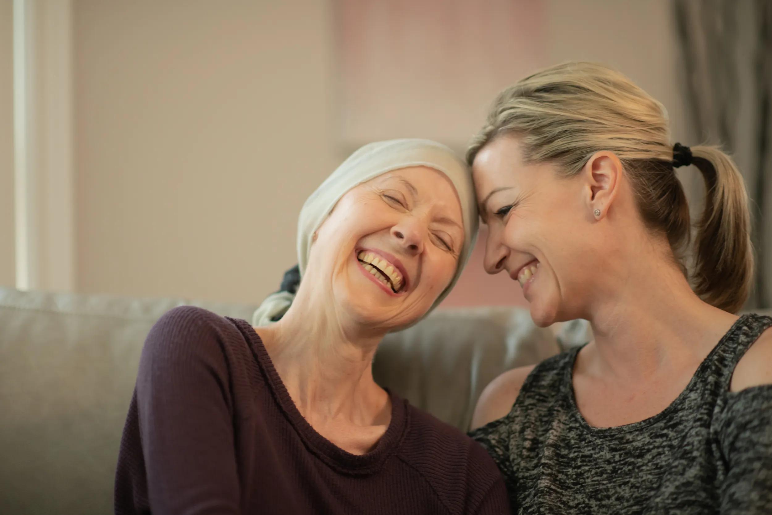 An elderly cancer patient sits on her sofa at home while she shares a laugh with her daughter.  They sit close, head to head.