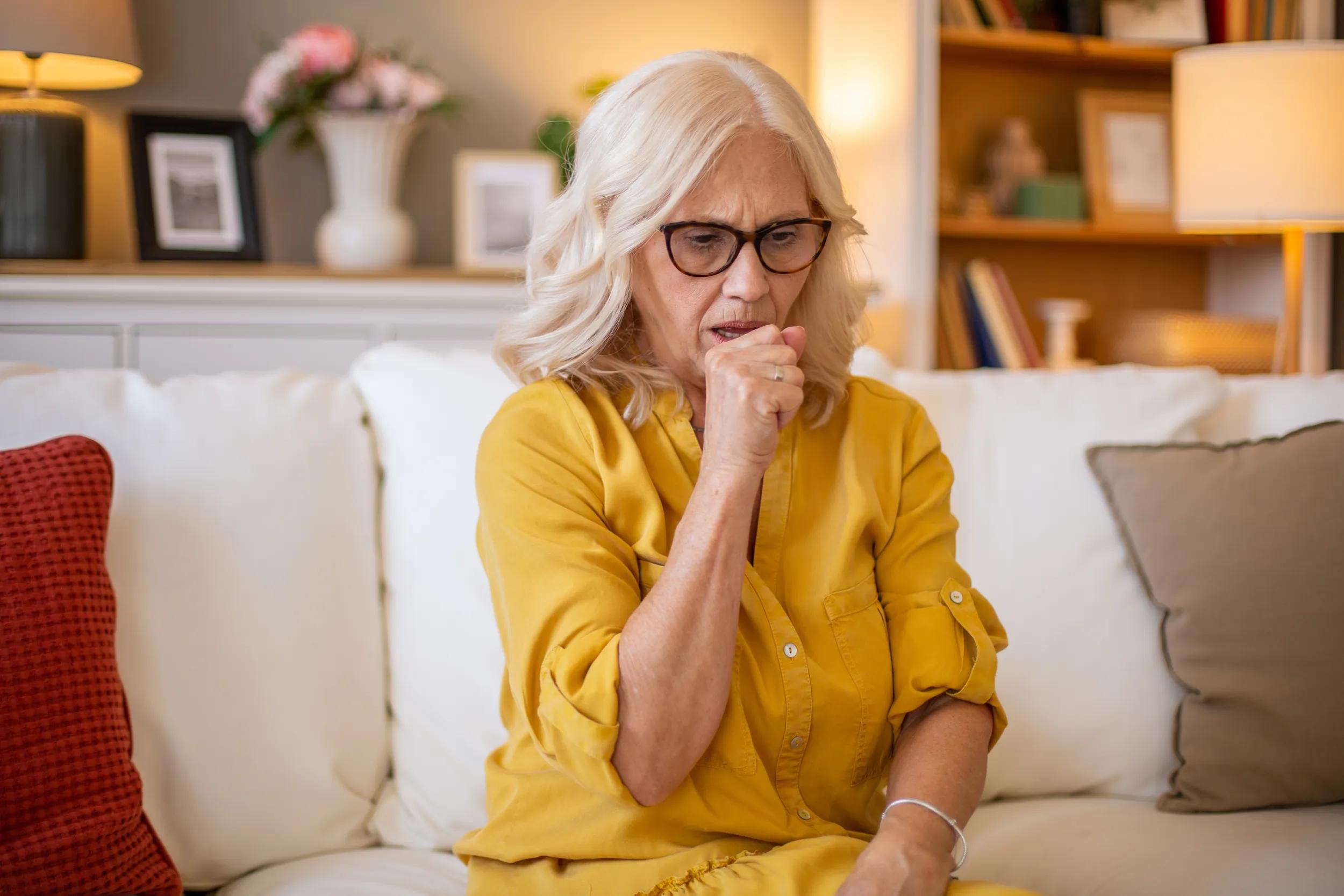 Senior woman coughing at home on sofa: feeling unwell