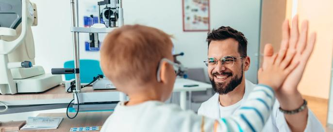 A smiling optometrist in a lab coat high-fives a young patient in a bright examination room, symbolizing trust and achievement during a medical visit.