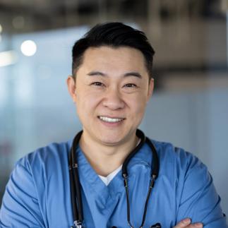 Close-up portrait of a young Asian male doctor standing in a blue uniform, arms crossed over his chest and looking at the camera