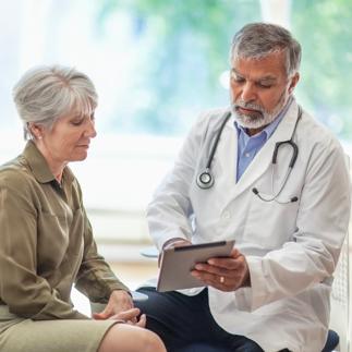 Asian Indian Pacific Islander senior man doctor medical professional talks using digital tablet to senior woman in health clinic