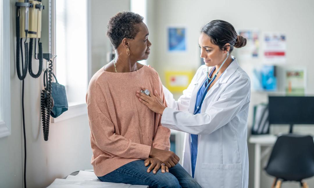A senior woman of African decent sits up on an exam table during a routine check-up.  She is sitting still and quiet as her female doctor listens to her heart.