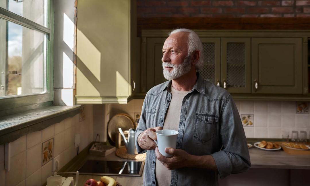 Senior man day dreaming while drinking coffee and looking through window at home.
