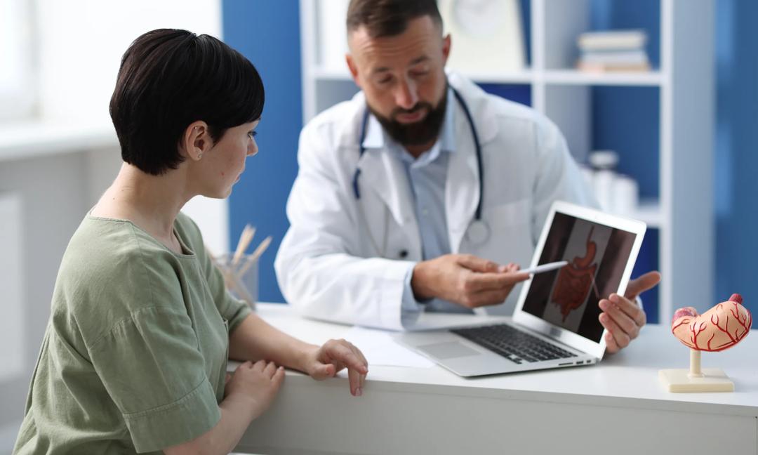 Gastroenterologist showing screen with illustration of large intestine to patient at table in clinic, selective focus