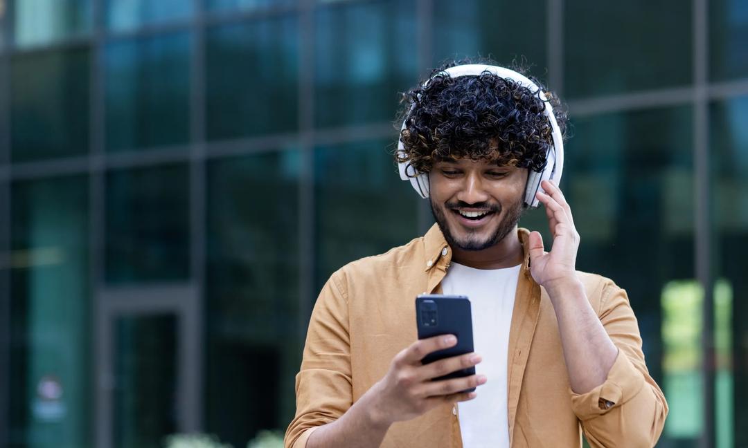 A young male Hispanic student is standing on the street near the campus wearing headphones with a phone in his hands. Listens to music, a podcast, talks on a video call