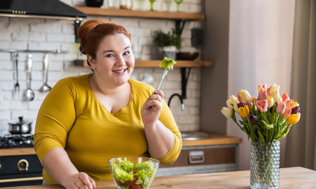 Obese young caucasian woman eating salad for healthy diet and eating habits in the kitchen. Chubby plus size woman having salad for breakfast