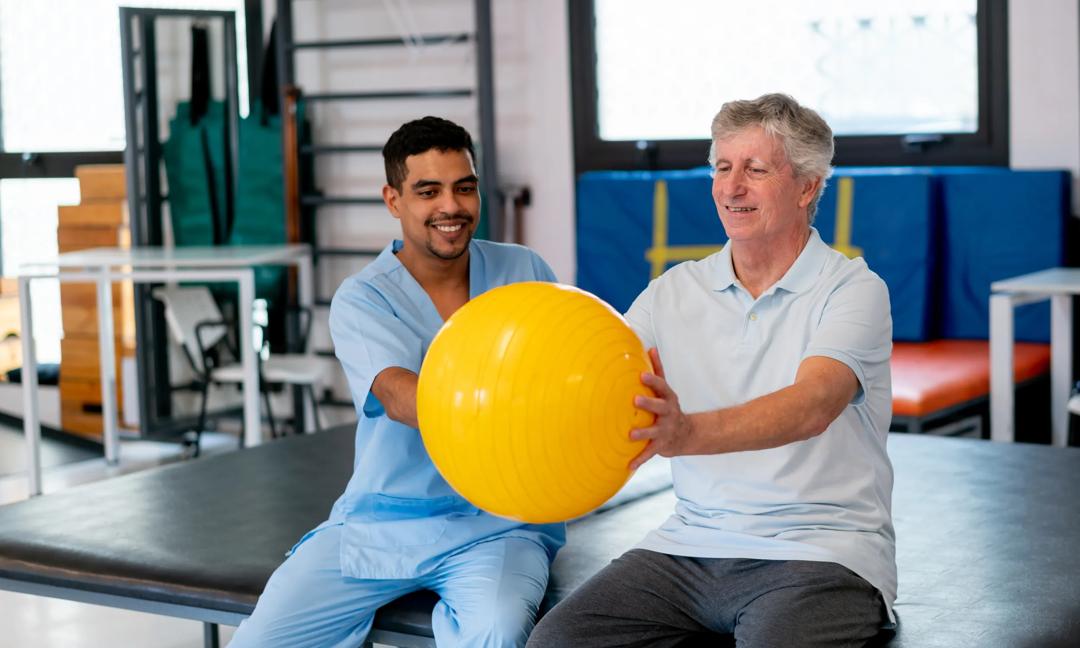 Caucasian senior male patient exercising his arms using a fitness ball and with the help of a male therapist at a physical recovery center