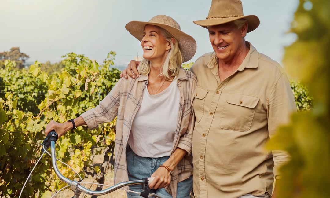 Smiling senior couple walking together while bonding on vineyard. Happy caucasian husband and wife pushing bicycle while enjoying day on farm after wine tasting on weekend. Man and woman on wine farm