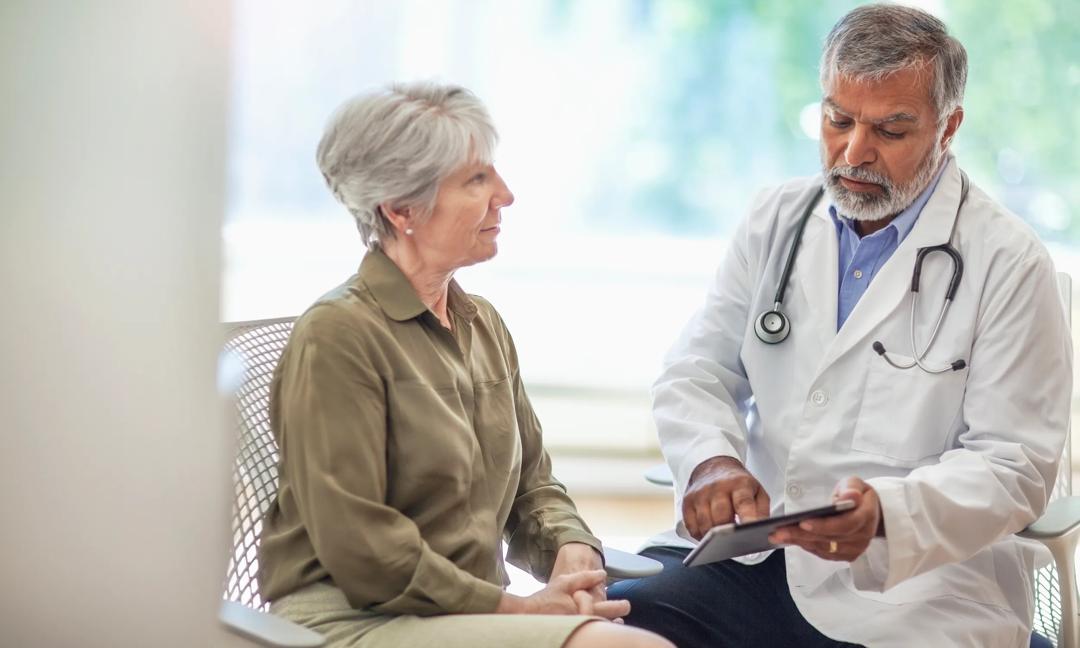 Asian Indian Pacific Islander senior man doctor medical professional talks using digital tablet to senior woman in health clinic