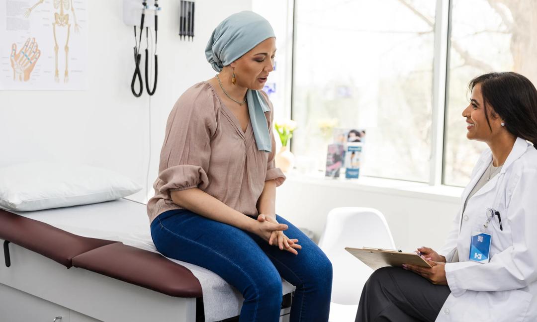 The mid adult female breast cancer survivor sits on the examination table and shares her symptoms as the mid adult female oncologist smiles encouragingly.
