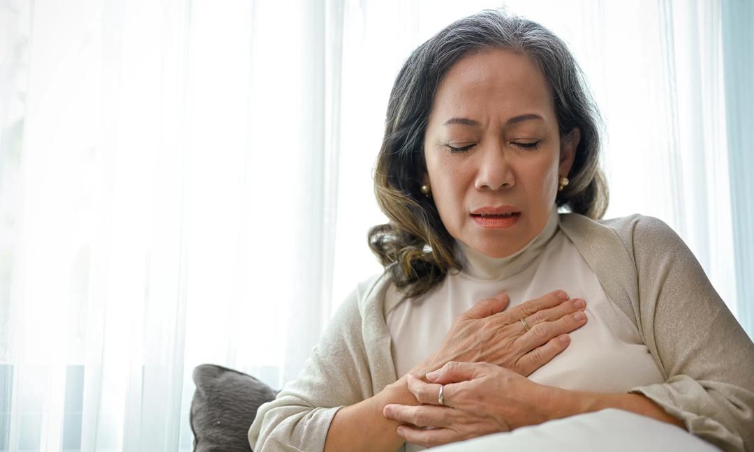 A sick Asian middle-aged woman with heart disease or heart ache, touching her chest and having a heart attack while relaxing at home. Health care concept