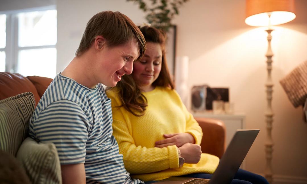 Young Downs Syndrome Couple Sitting On Sofa Using Laptop At Home