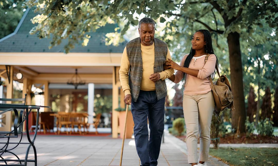 Black senior man with walking cane and his daughter taking a walk through the park of a nursing home.