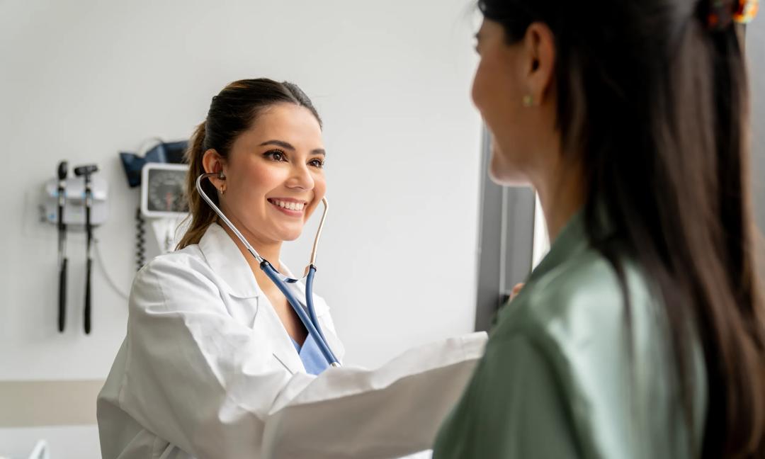 Happy Latin American doctor listening to the heartbeat of a patient at the hospital and smiling - medical exam concepts