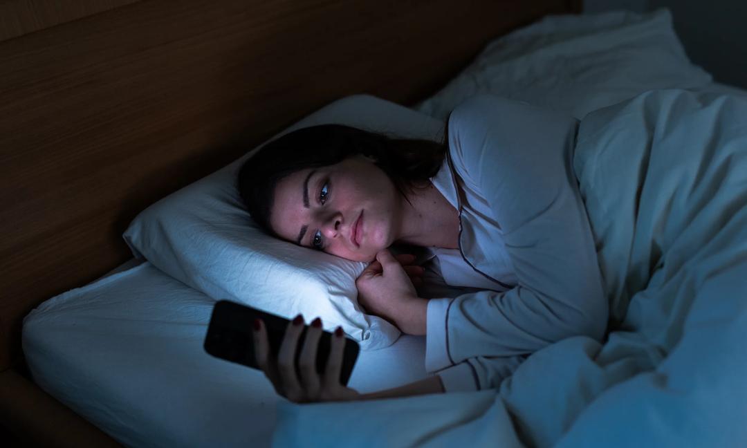 Young Caucasian woman using smartphone lying in bed at night. Insomnia and social media addiction. Copy space