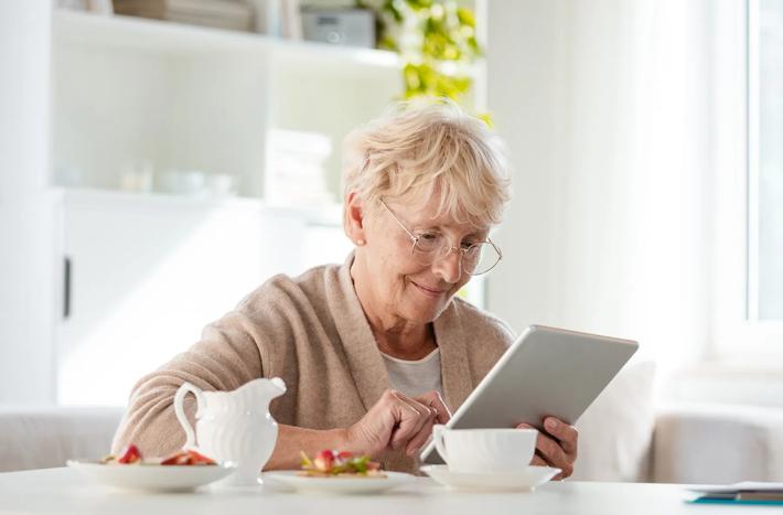 Smiling senior woman sitting at the table in her house alone and reading e-book.