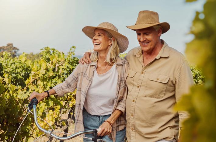 Smiling senior couple walking together while bonding on vineyard. Happy caucasian husband and wife pushing bicycle while enjoying day on farm after wine tasting on weekend. Man and woman on wine farm