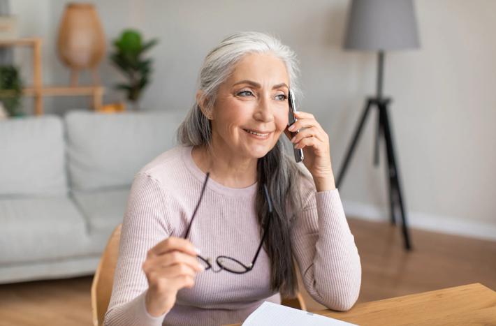 mature gray-haired lady hold glasses speaks by phone with client in living room interior, copy space
