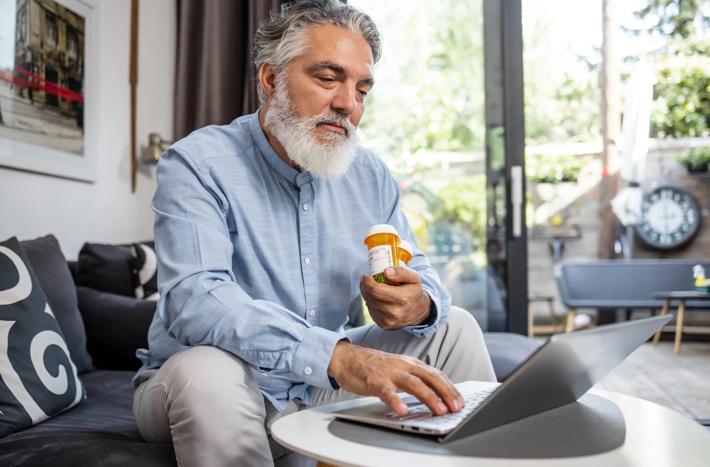Photo of a senior man at home holding a pill bottle and using laptop. Health technology concept.