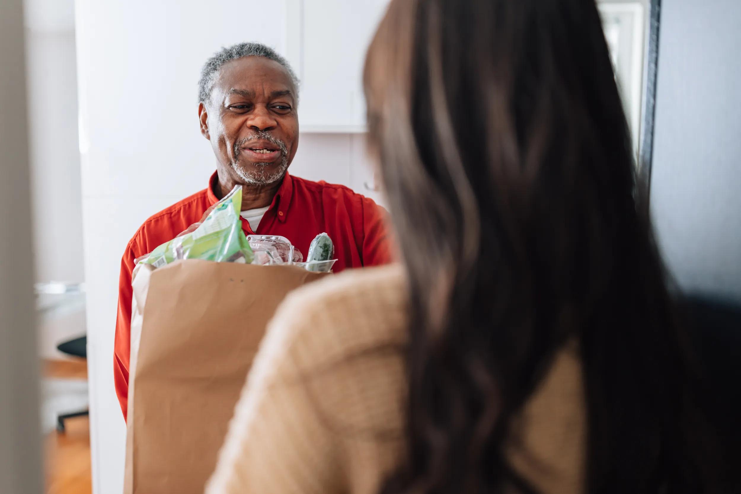 A young female Caucasian volunteer brings bag with food to an older man's front door